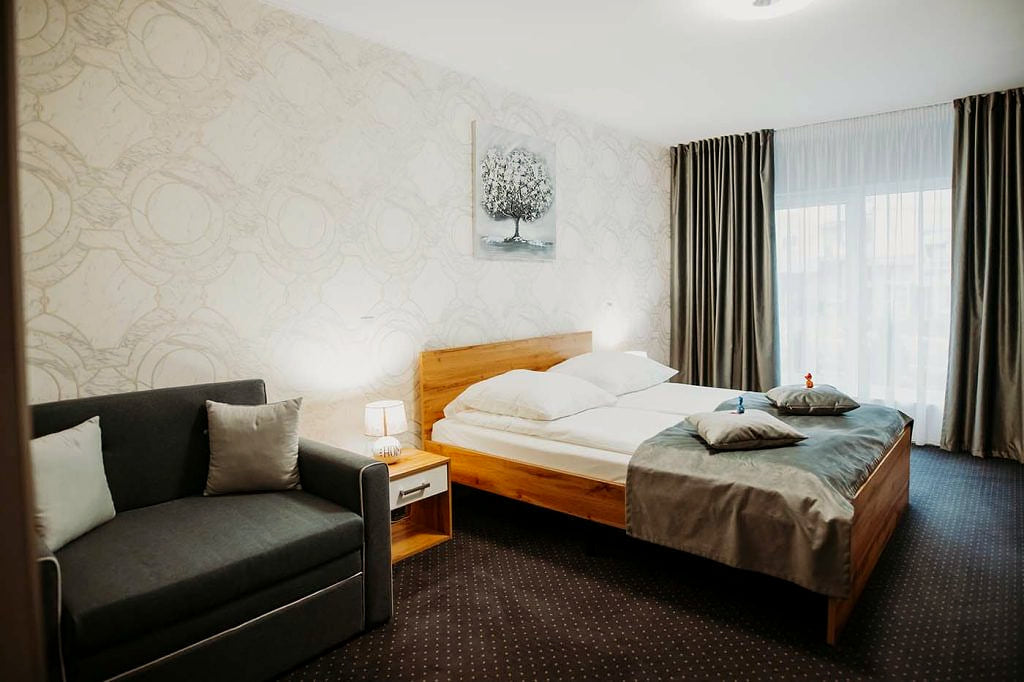 A room at the Epic Centrum Hotel with a wooden bed, a grey sofa, patterned wallpaper and long curtains at the window.