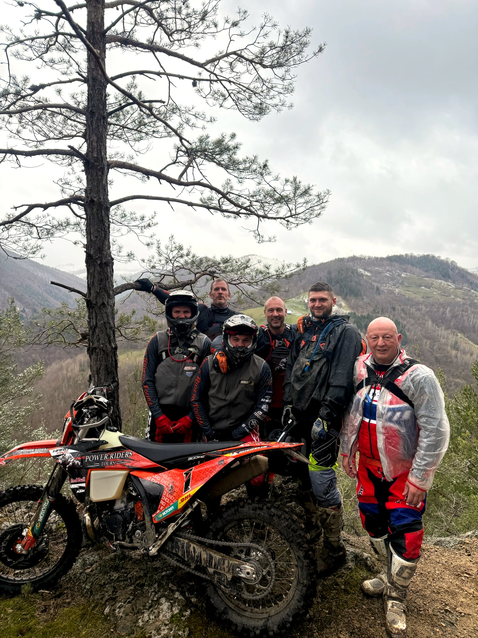 Five riders beside a pine tree on a hilltop, an orange KTM in front of them