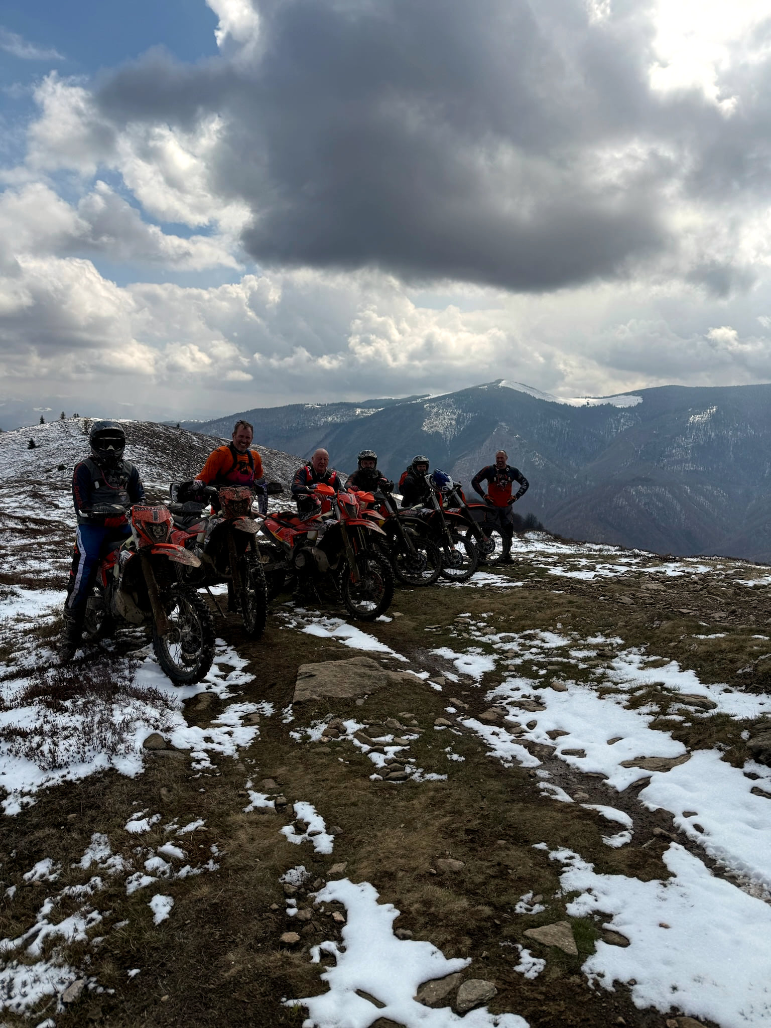 A group of riders with their bikes on a mountain ridge with patches of snow under a cloudy sky