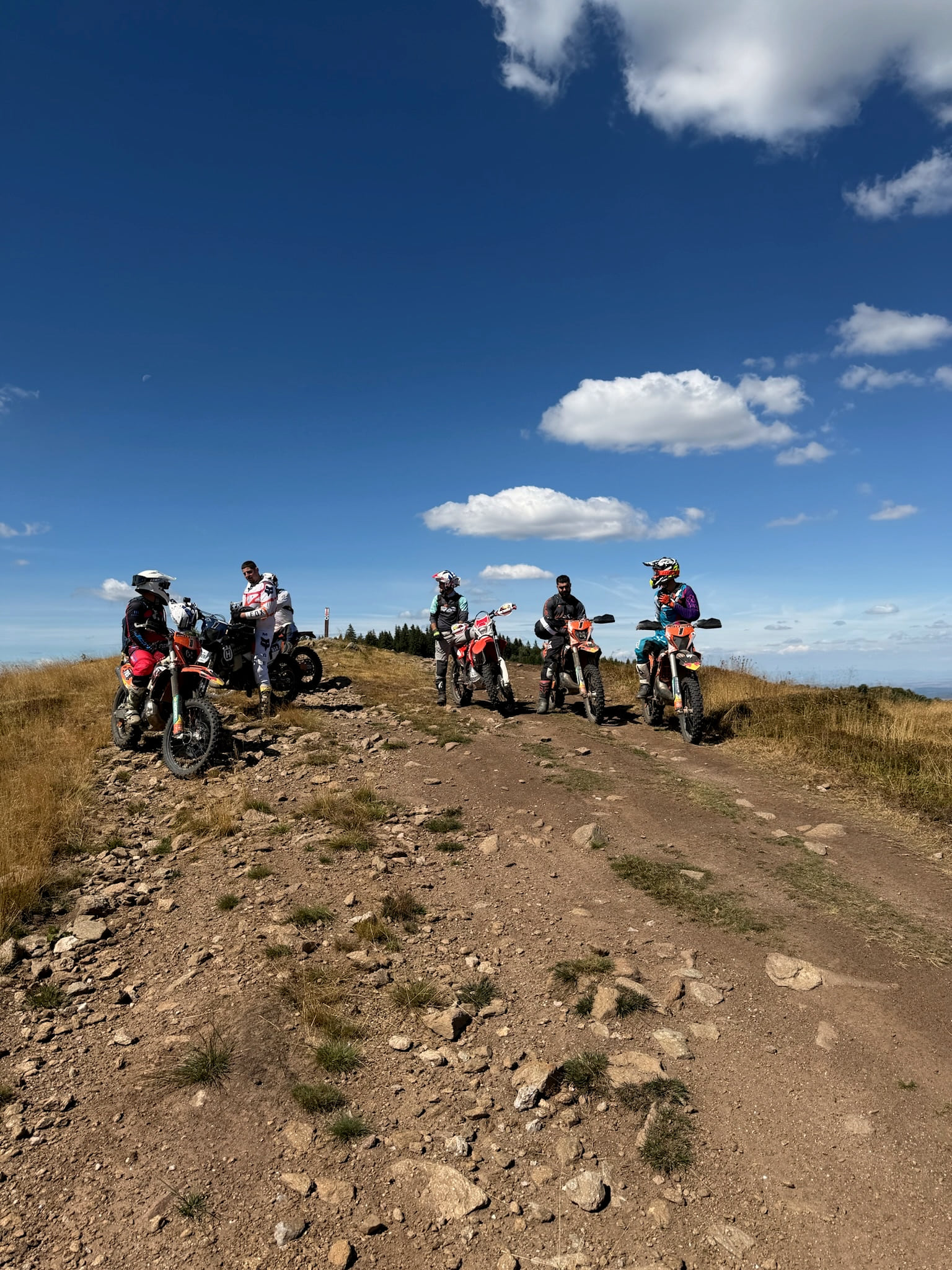 Riders and their bikes in a line along a high ridge under a blue sky with white clouds