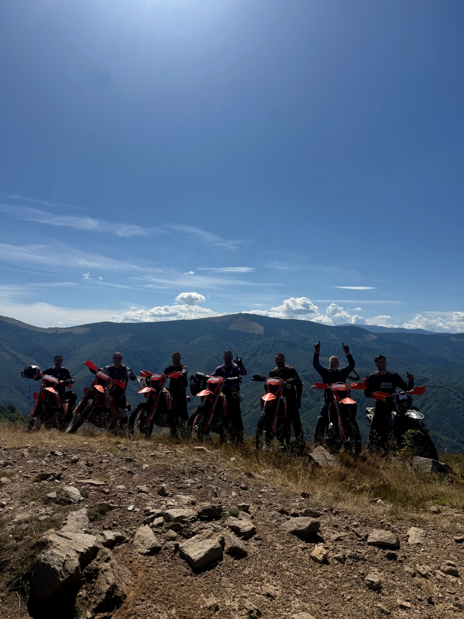 Seven riders with their bikes lined up on a rocky ridge under a clear blue sky