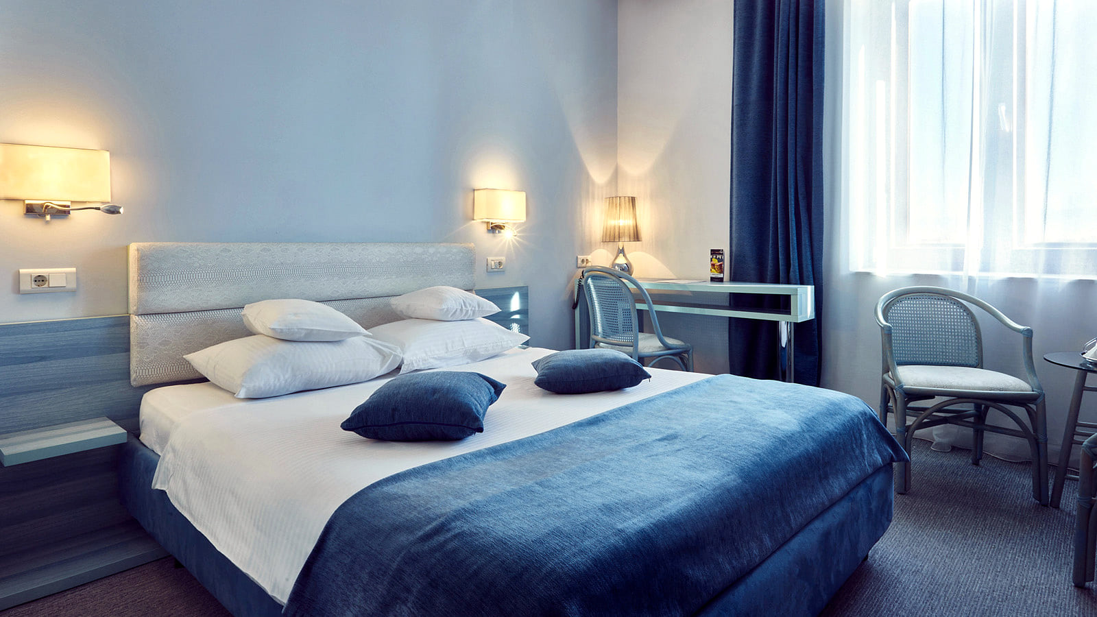 A room at the Continental Forum Sibiu with a wide bed in blue and white, two wall lamps and a desk by the window.