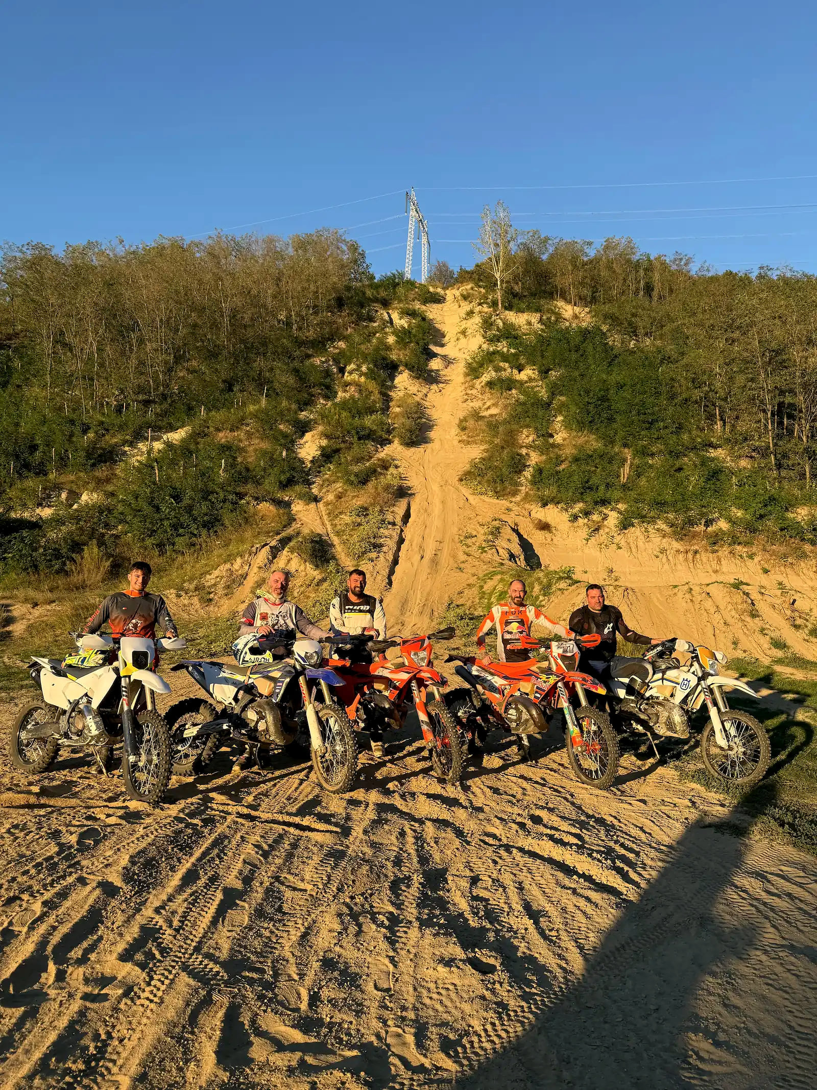 Five riders stand with their enduro bikes lined up on the sand floor of a quarry at golden hour, a steep hill climb rising behind them.