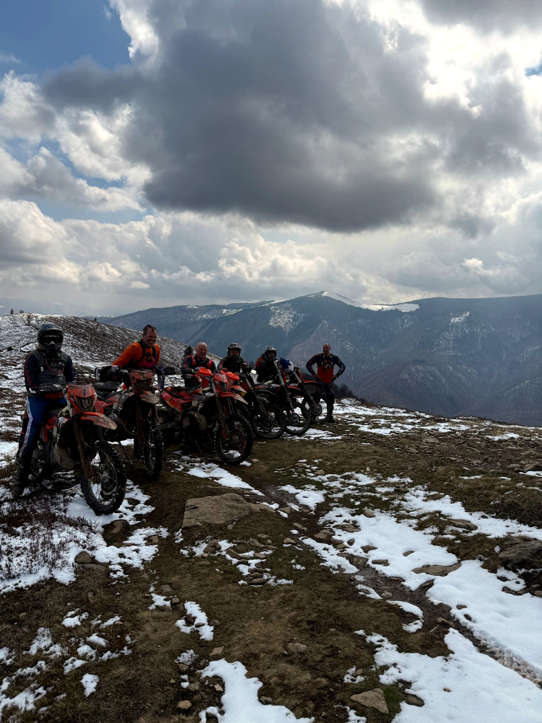 Group of six riders with their bikes on a ridge with patches of snow in the mountains