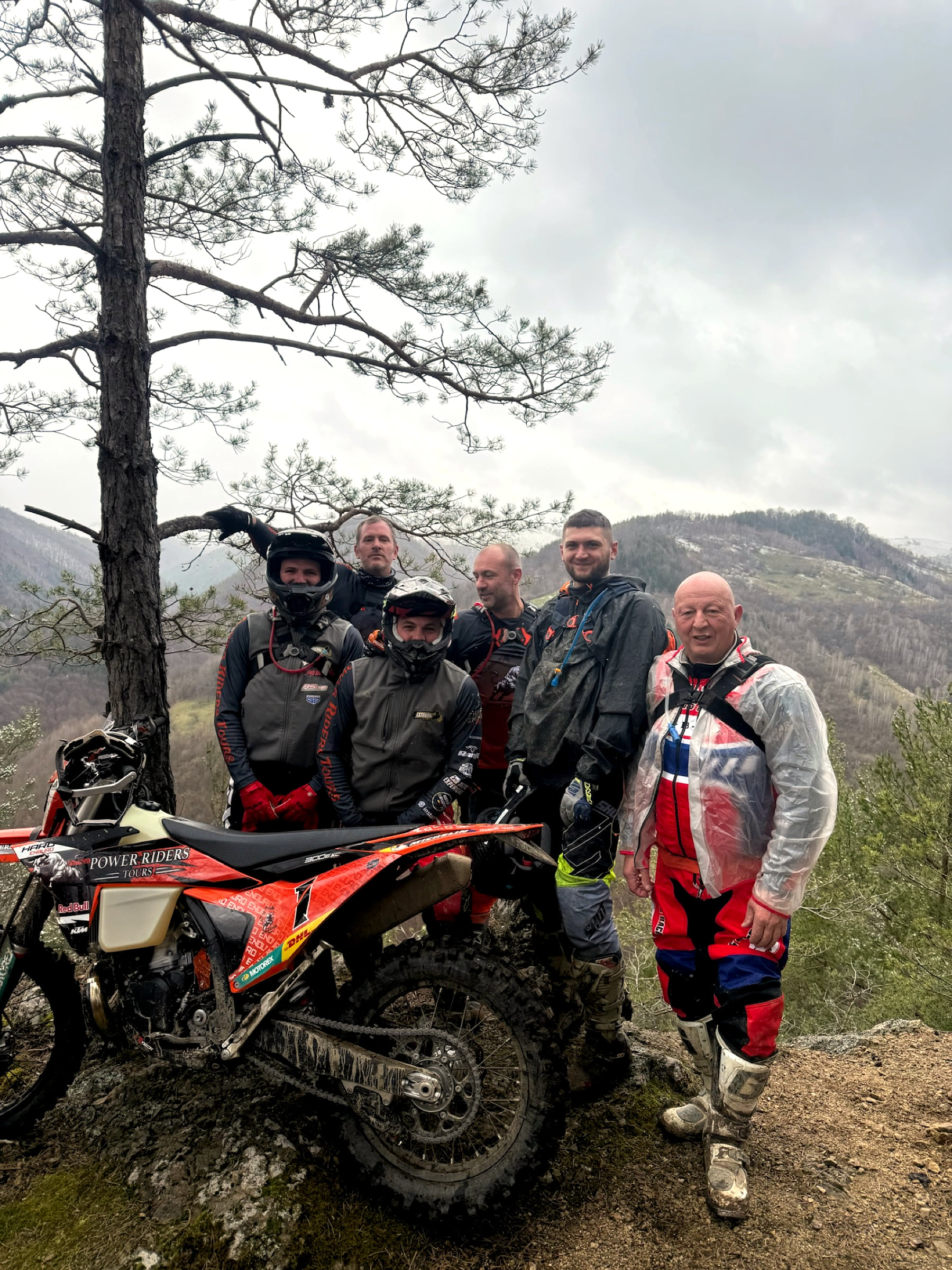 Three riders in muddy gear around a KTM on a high pasture in the mist