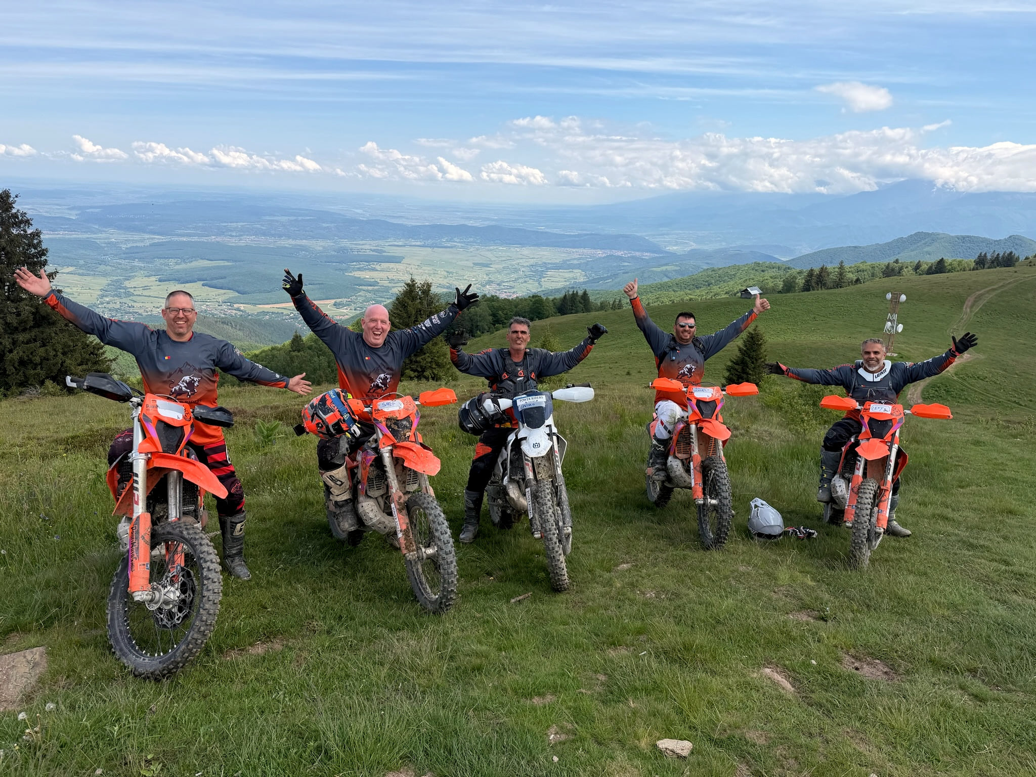 Five riders on orange KTM and white enduro bikes on a high meadow, arms raised, a wide valley below