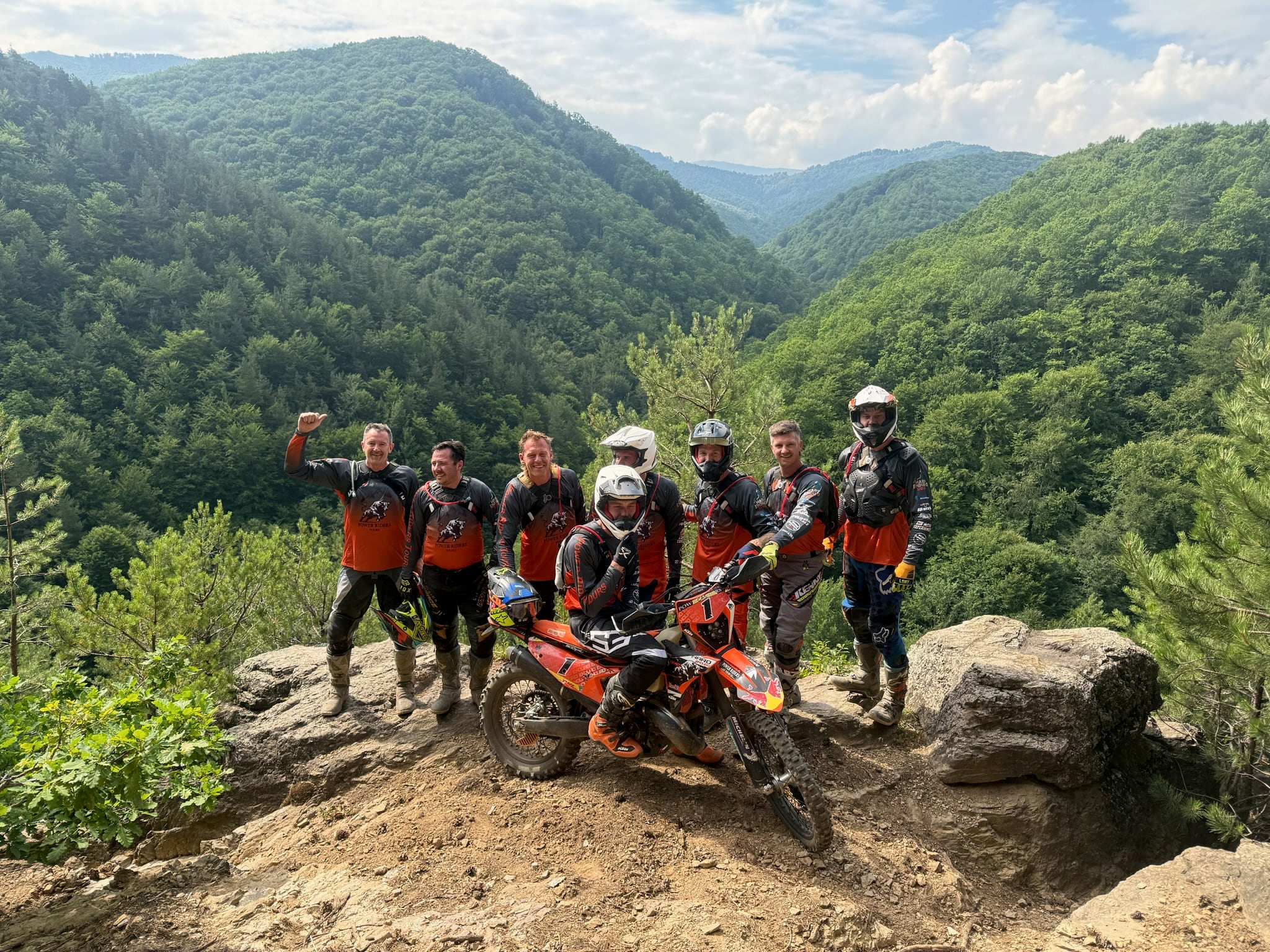 Eight riders in orange Power Riders Tours shirts on a rock outcrop above a deep forested valley, a KTM in the middle