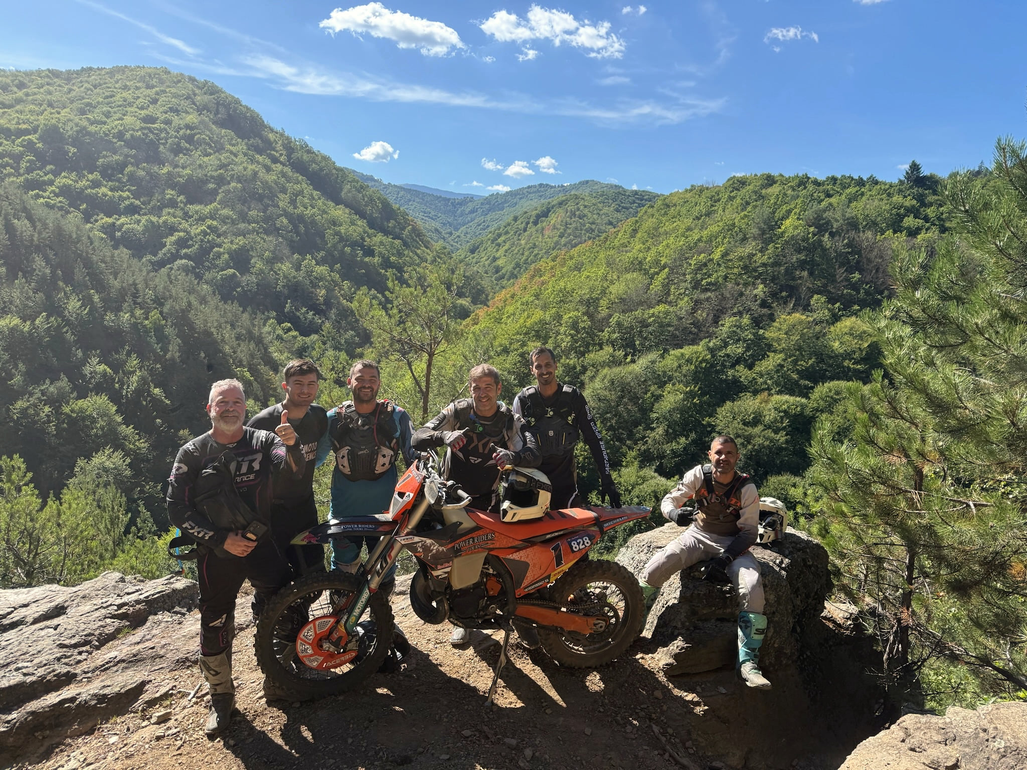 A group of riders with their KTM bikes on a sunny rock outcrop above green mountain valleys