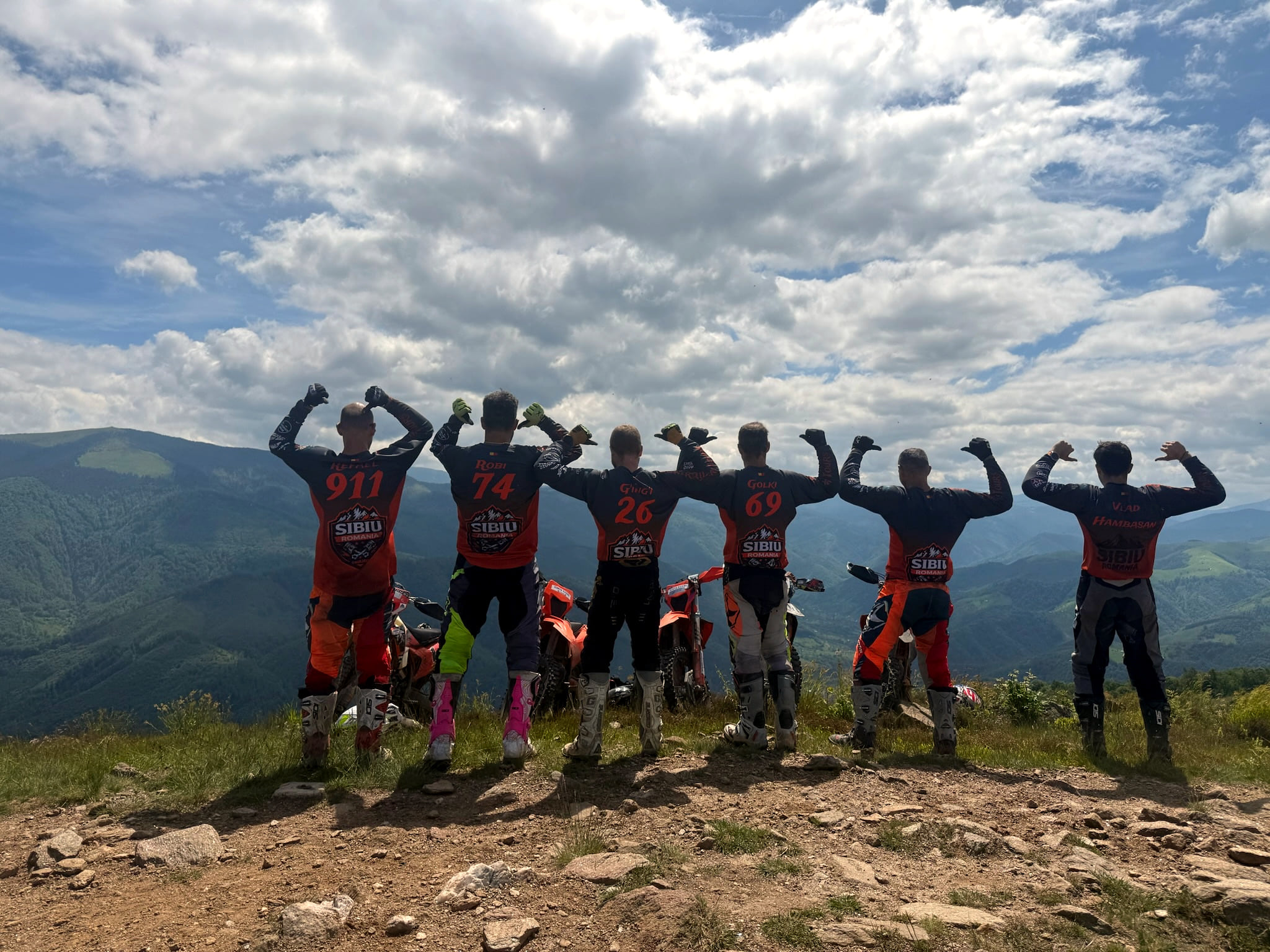 Seven riders in numbered Sibiu jerseys seen from behind, flexing their arms on a summit above the mountains