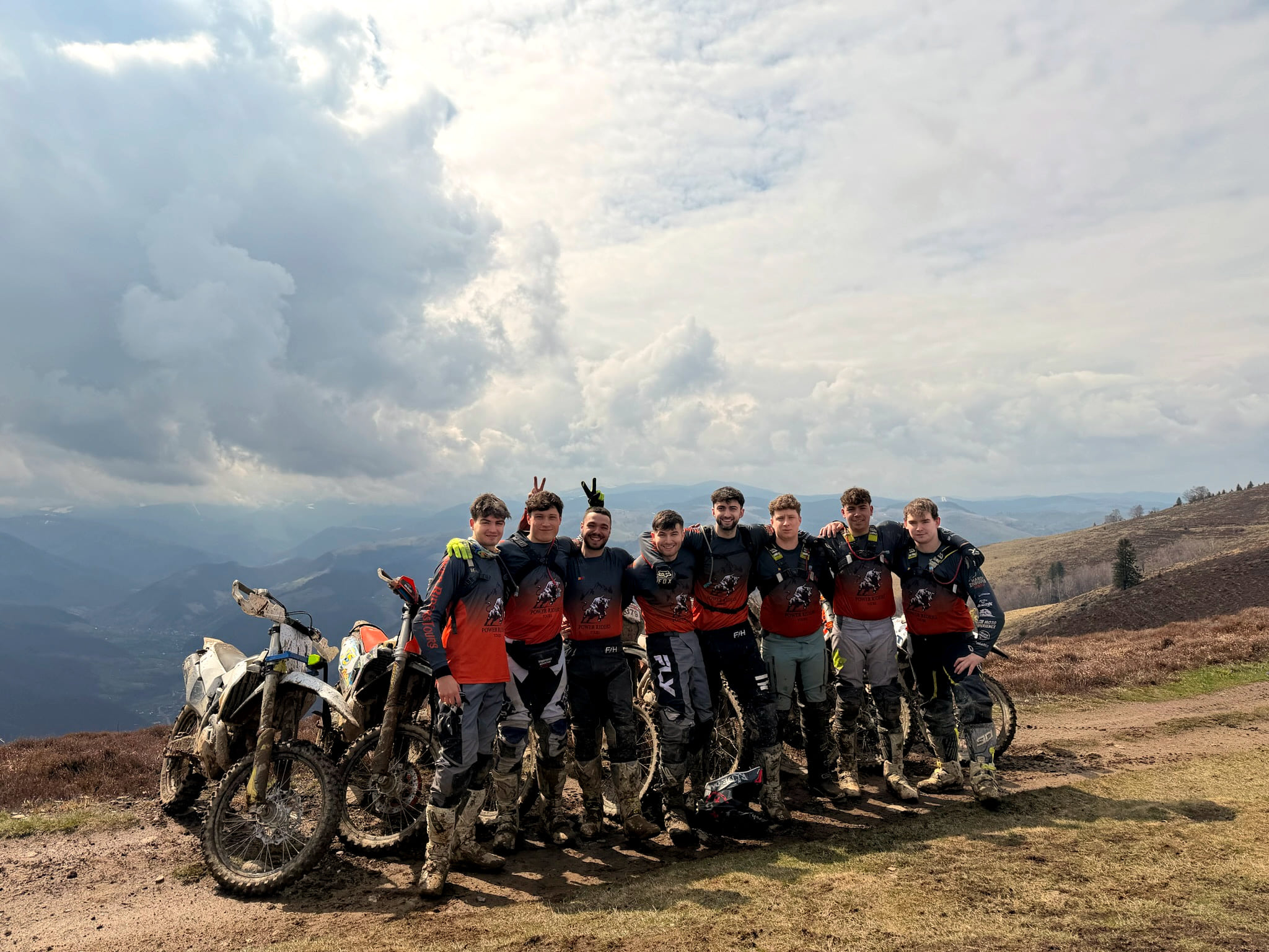 A group of riders stand in a row behind their bikes on a ridge under a wide, cloudy sky.