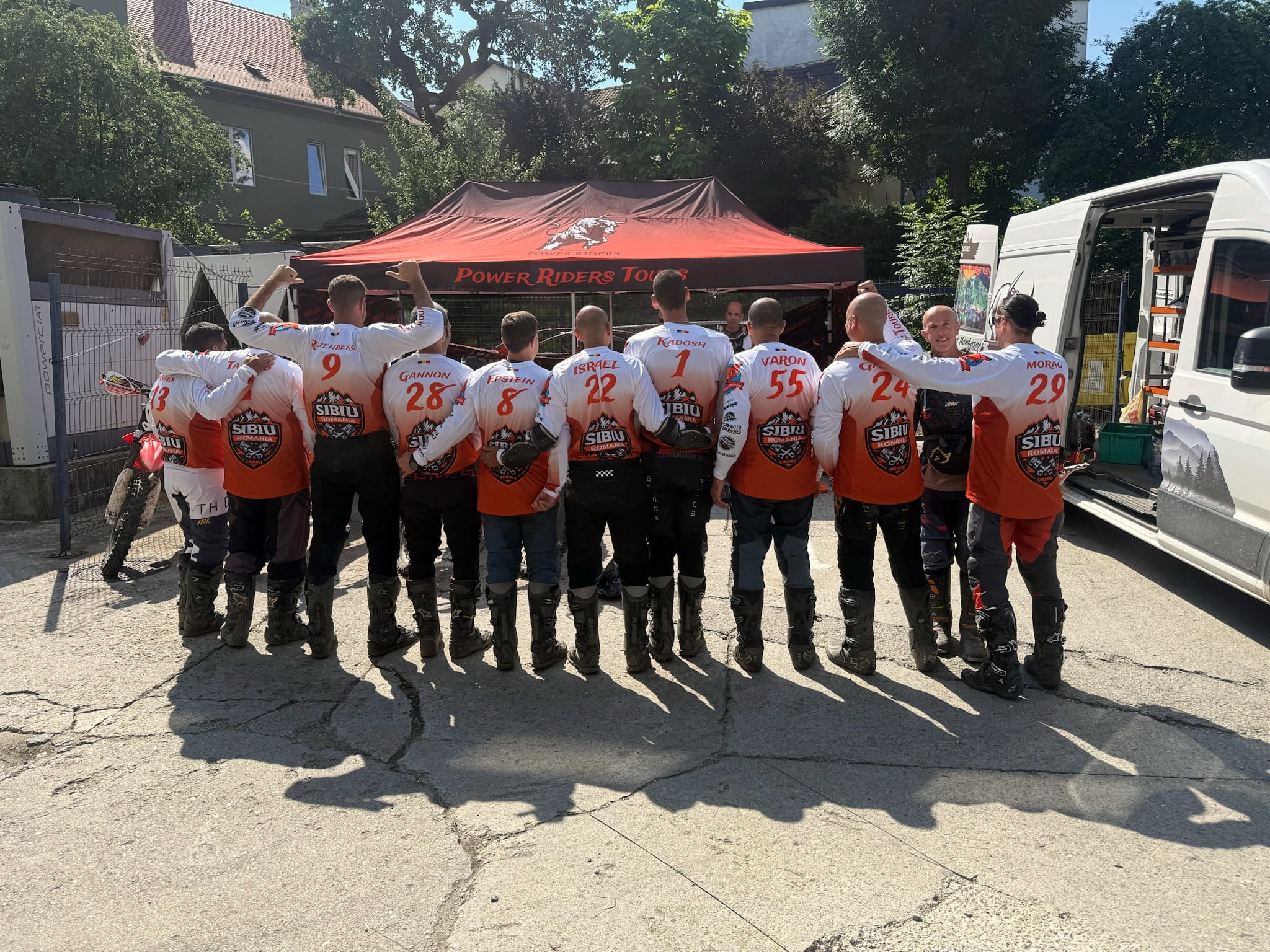 Fourteen riders in white and orange numbered Sibiu jerseys, arms round each other, in front of the Power Riders Tours tent and van