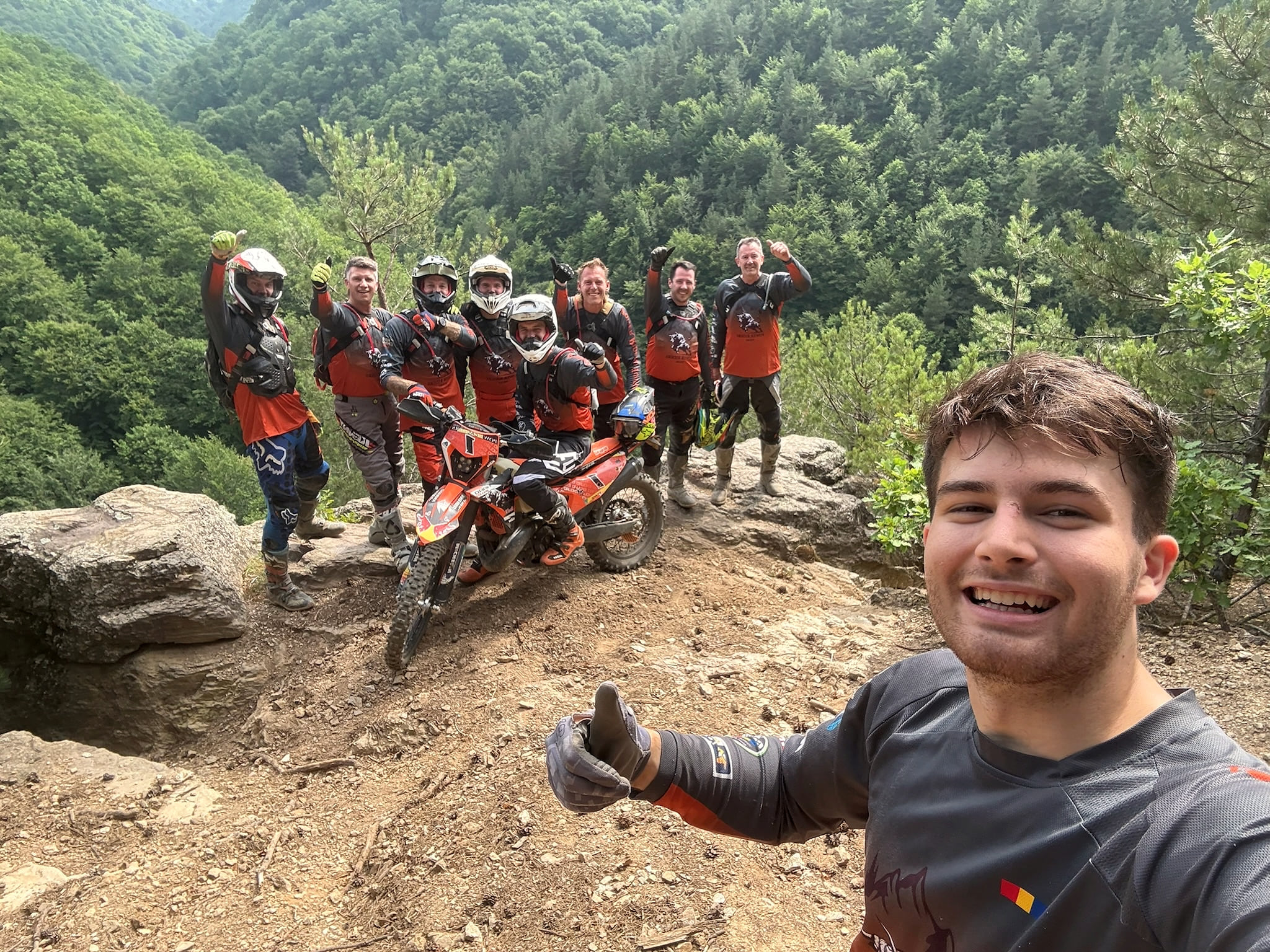 A rider takes a selfie in front of his group of eight on a rock outcrop above a forested valley