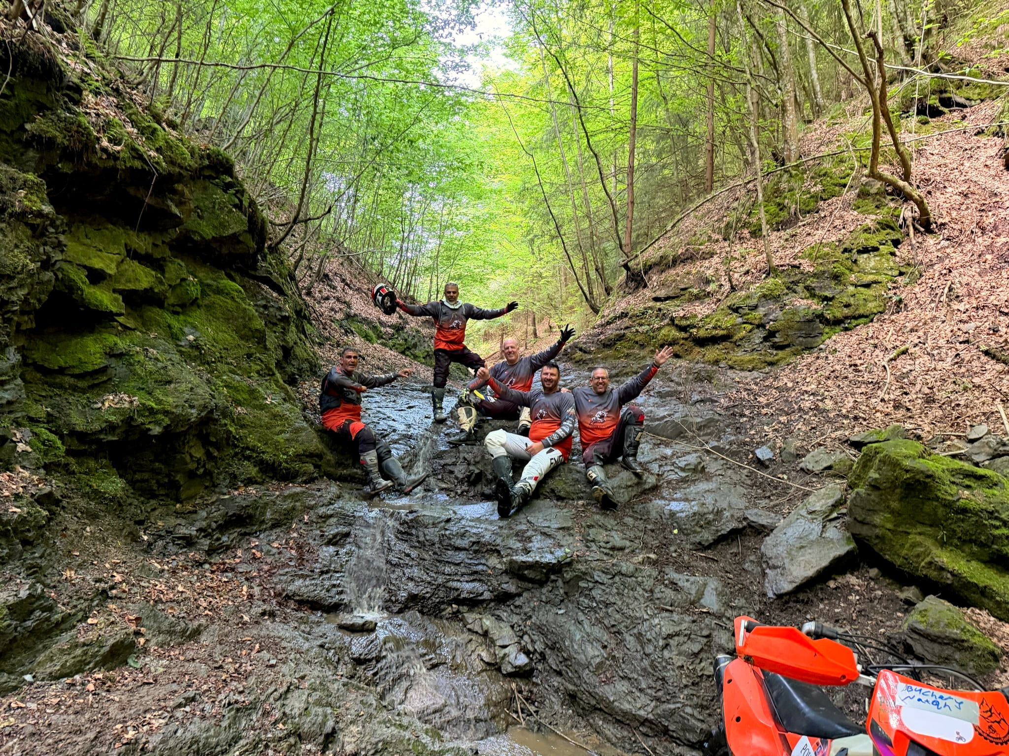 Five riders cheering in a rocky forest creek, their orange KTM bikes nearby