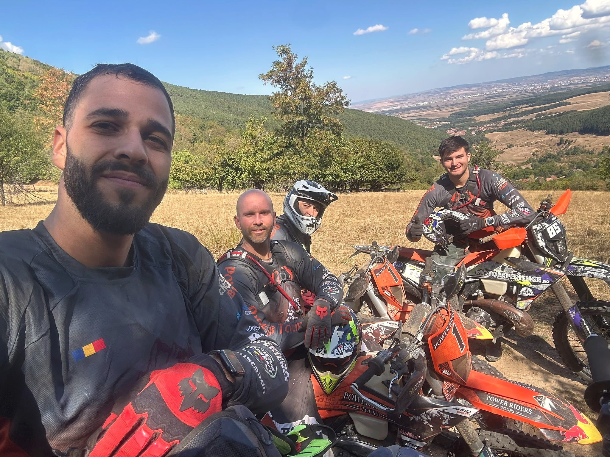 Four smiling riders take a selfie on a dry ridge with their orange bikes, a wide plain behind