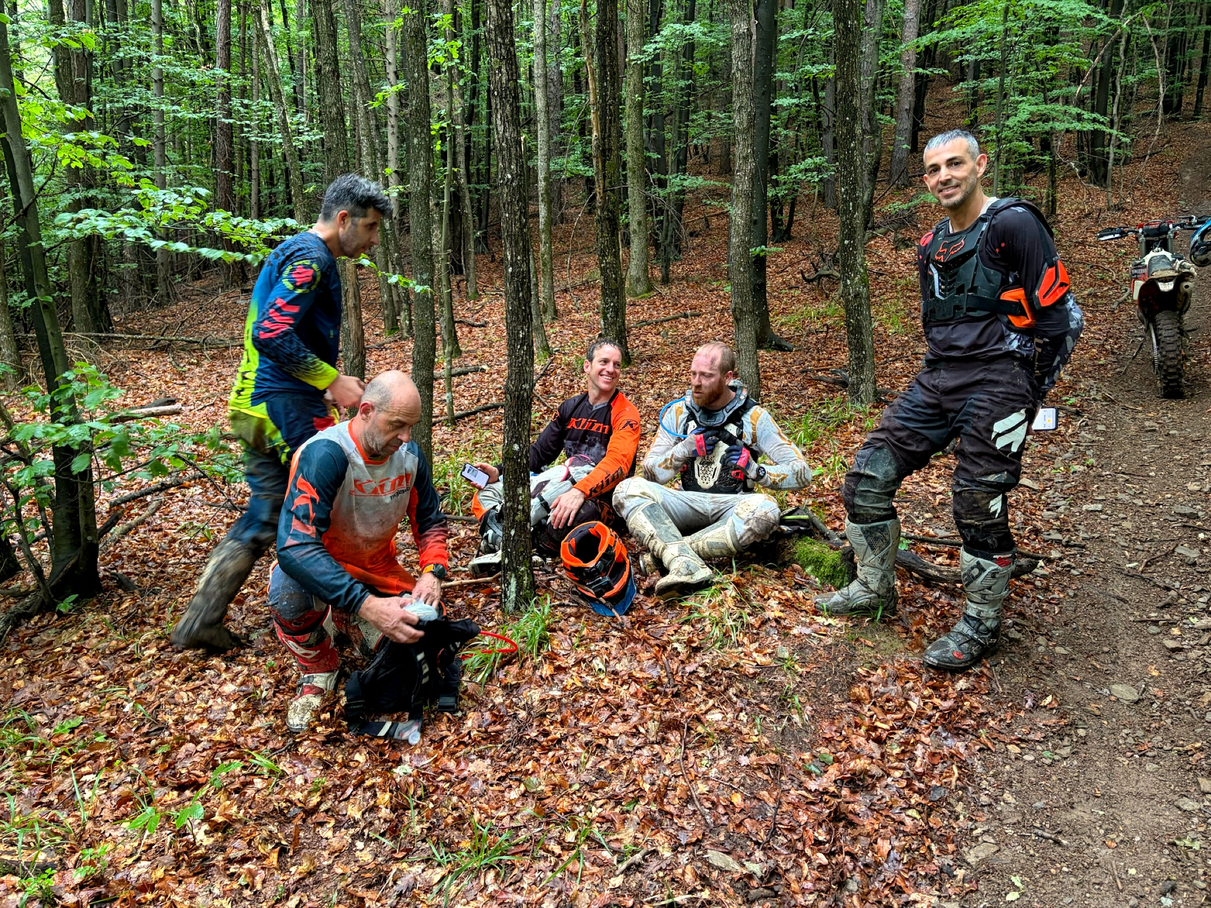 Riders resting on the forest floor beside their bikes, helmets off, during a riding day