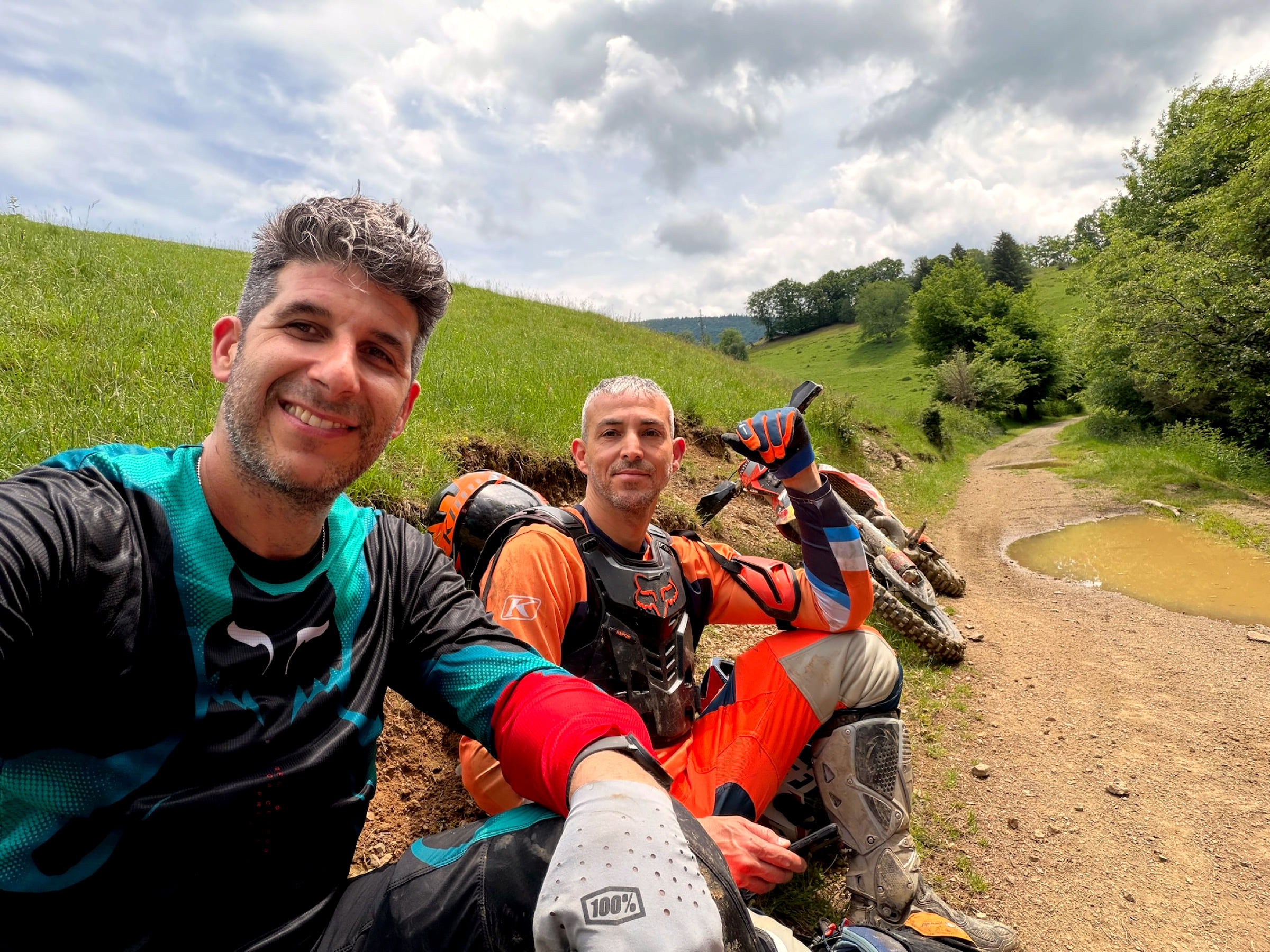 Two riders sitting on a grass bank beside a dirt track, green hills behind