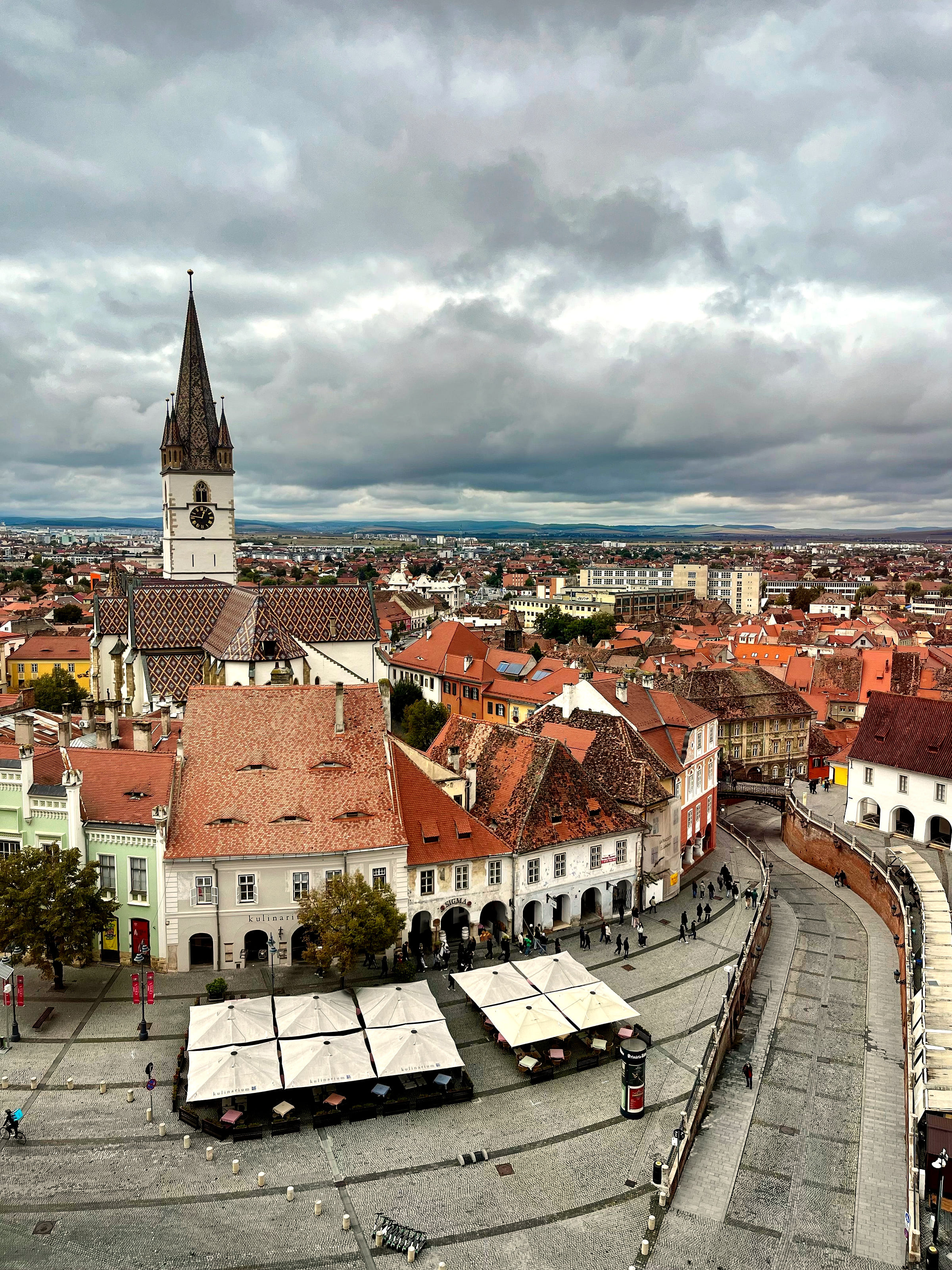 Sibiu's old town from above: café umbrellas on a cobbled square, red roofs and the Lutheran Cathedral's tower.