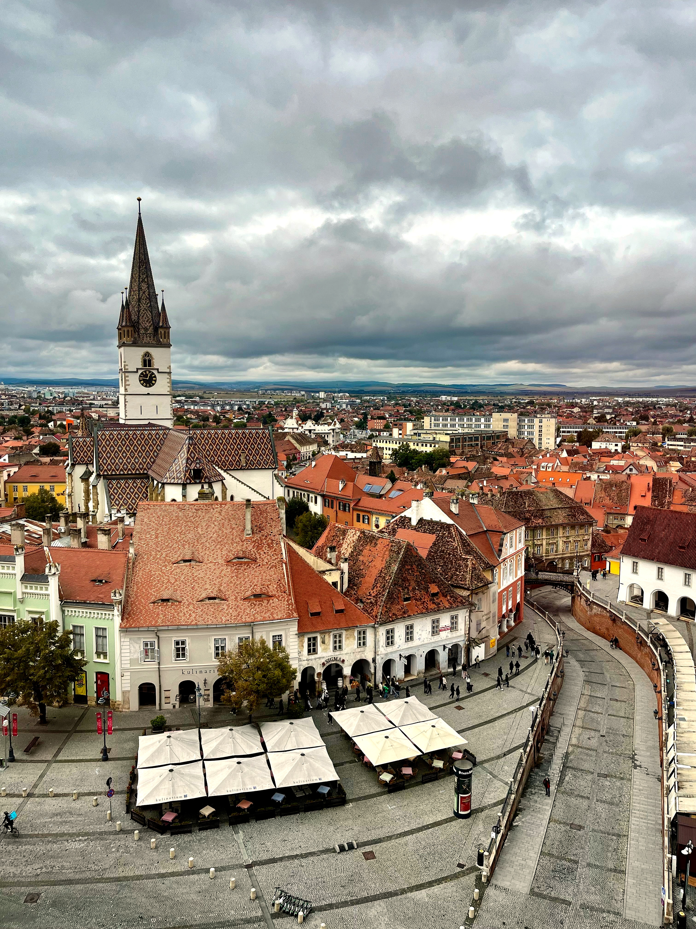 Sibiu's old town from above: café umbrellas on a cobbled square, red roofs and the Lutheran Cathedral's tower.
