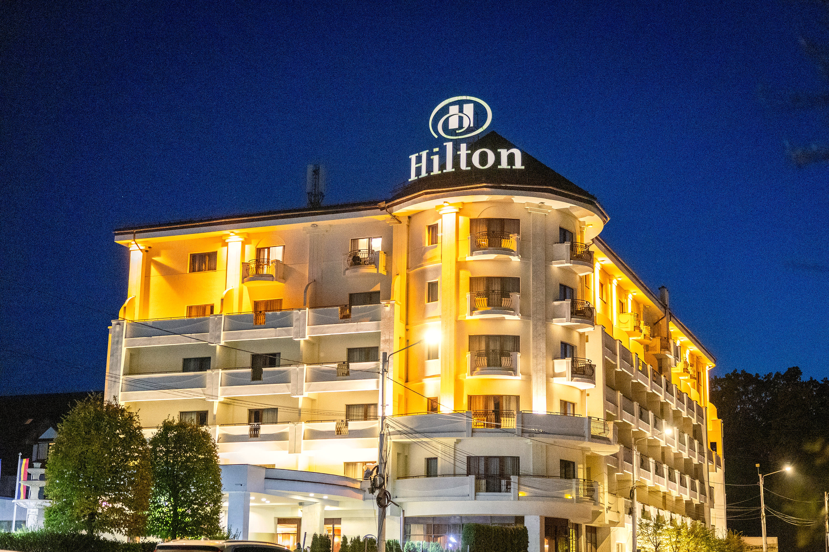 Hilton Sibiu at night: the lit cream hotel with the Hilton sign on its rounded corner tower, trees around it.