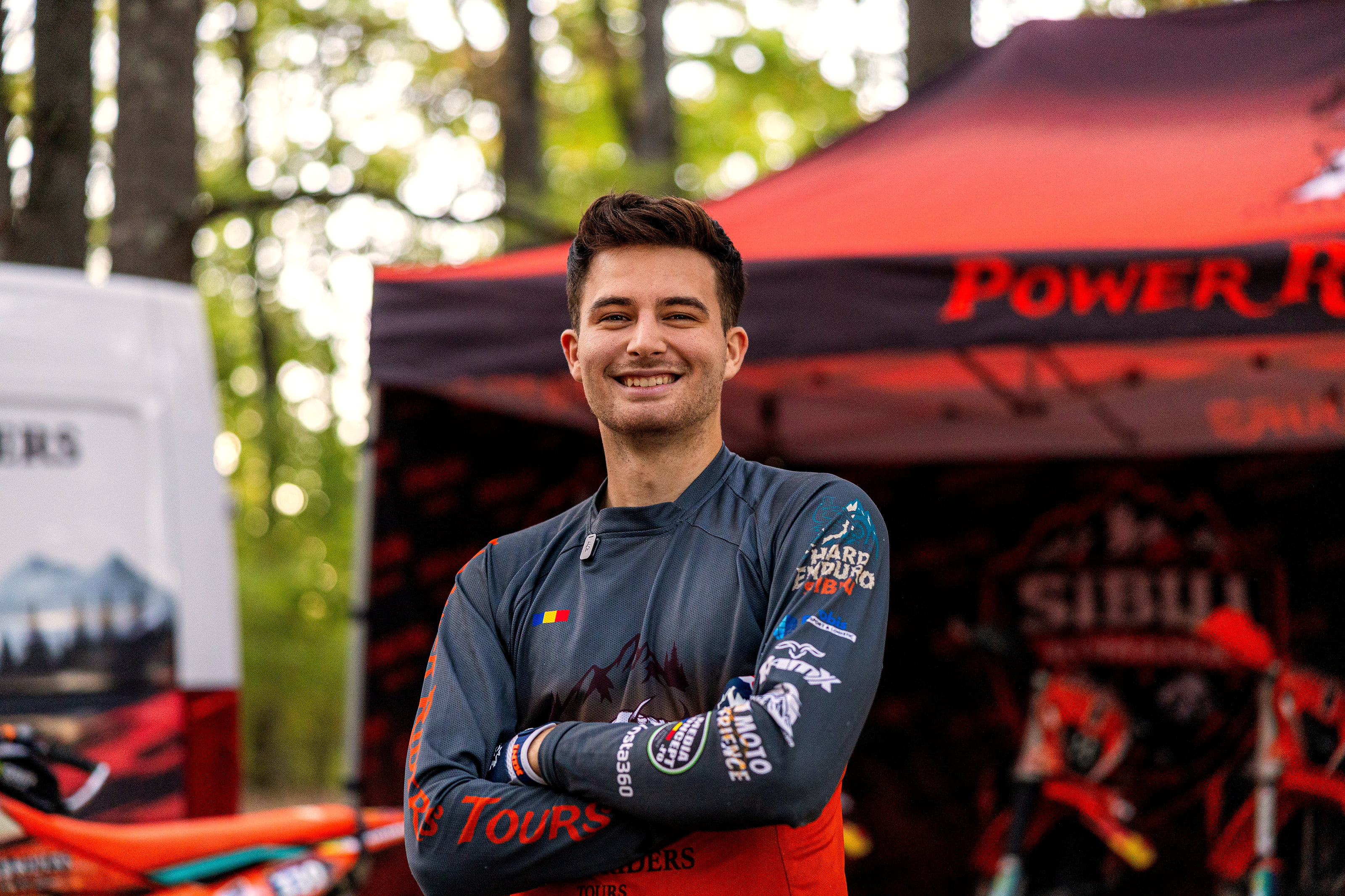 Vlad Hambasan, lead guide at Power Riders Tours, smiling with arms crossed in front of the team's tent and bikes in the forest.