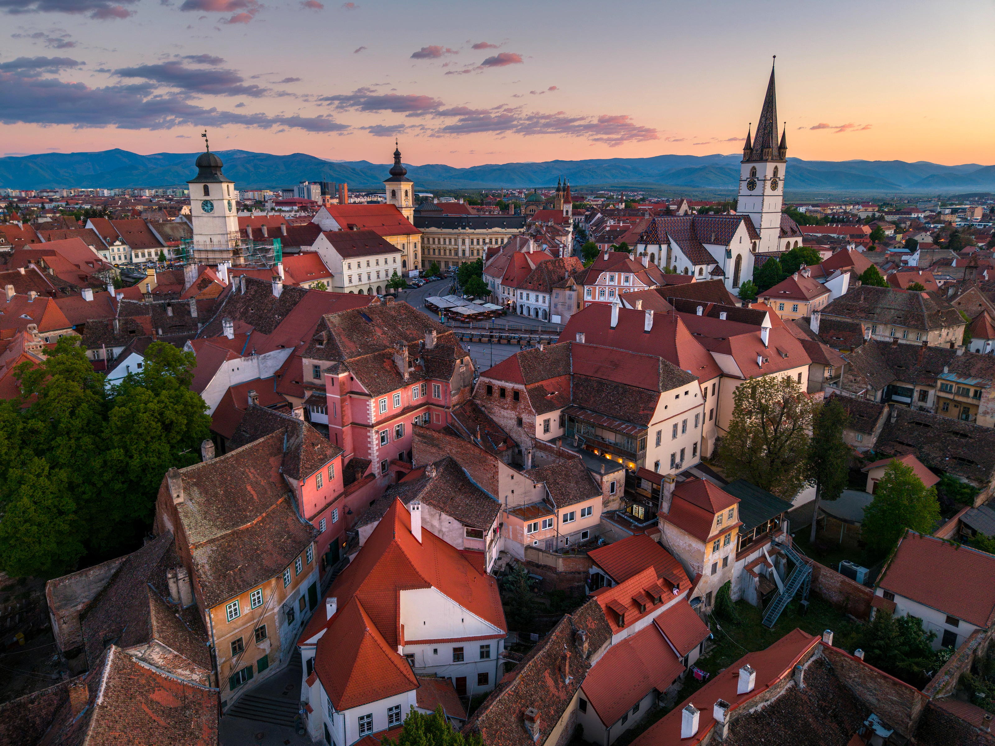 Die Altstadt von Sibiu aus der Luft in der Dämmerung: rote Dächer, alte Türme und die Stadtpfarrkirche, dahinter Berge.