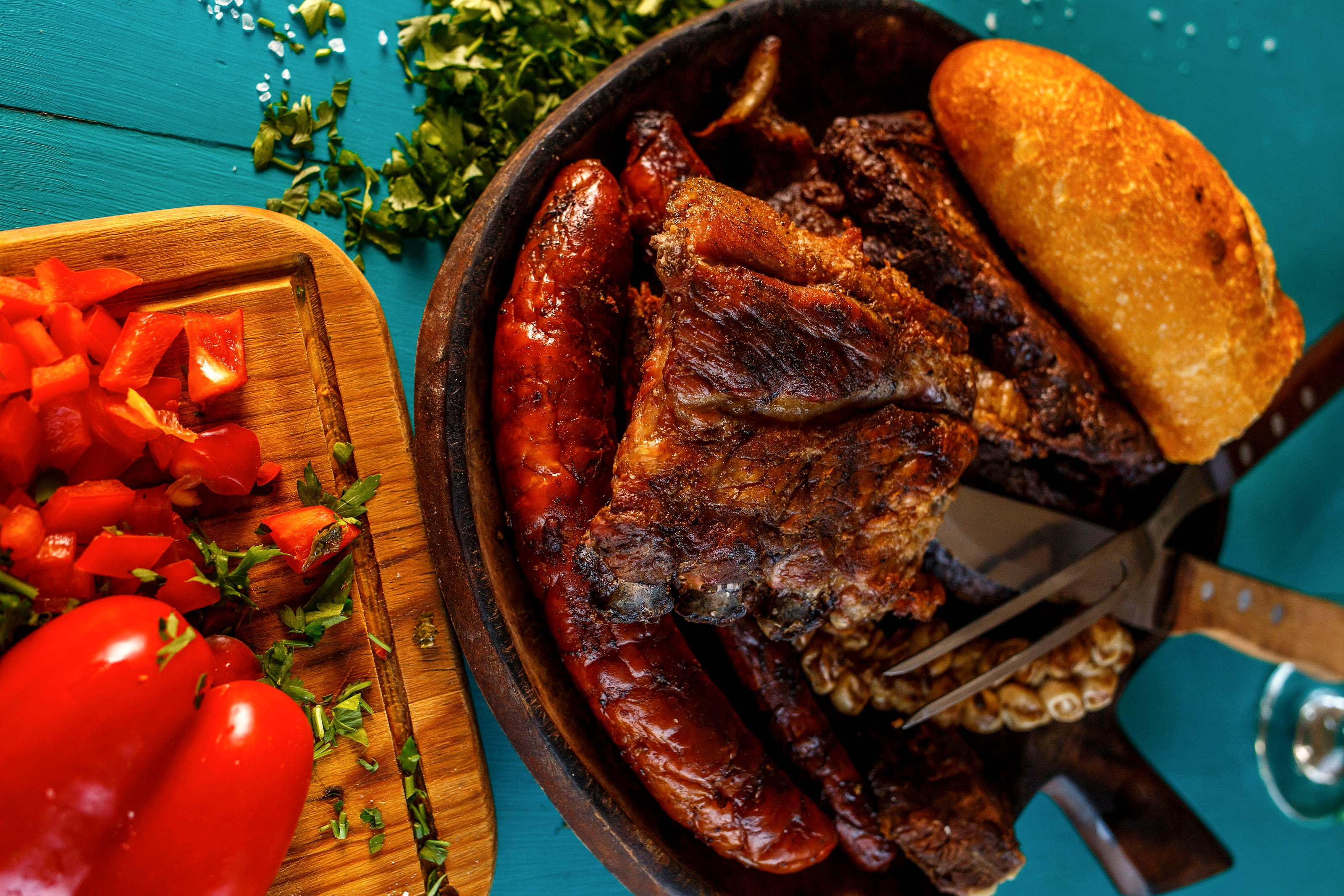Grilled sausages and meat in an iron pan with a bread roll, beside red peppers and chopped tomatoes on a board.